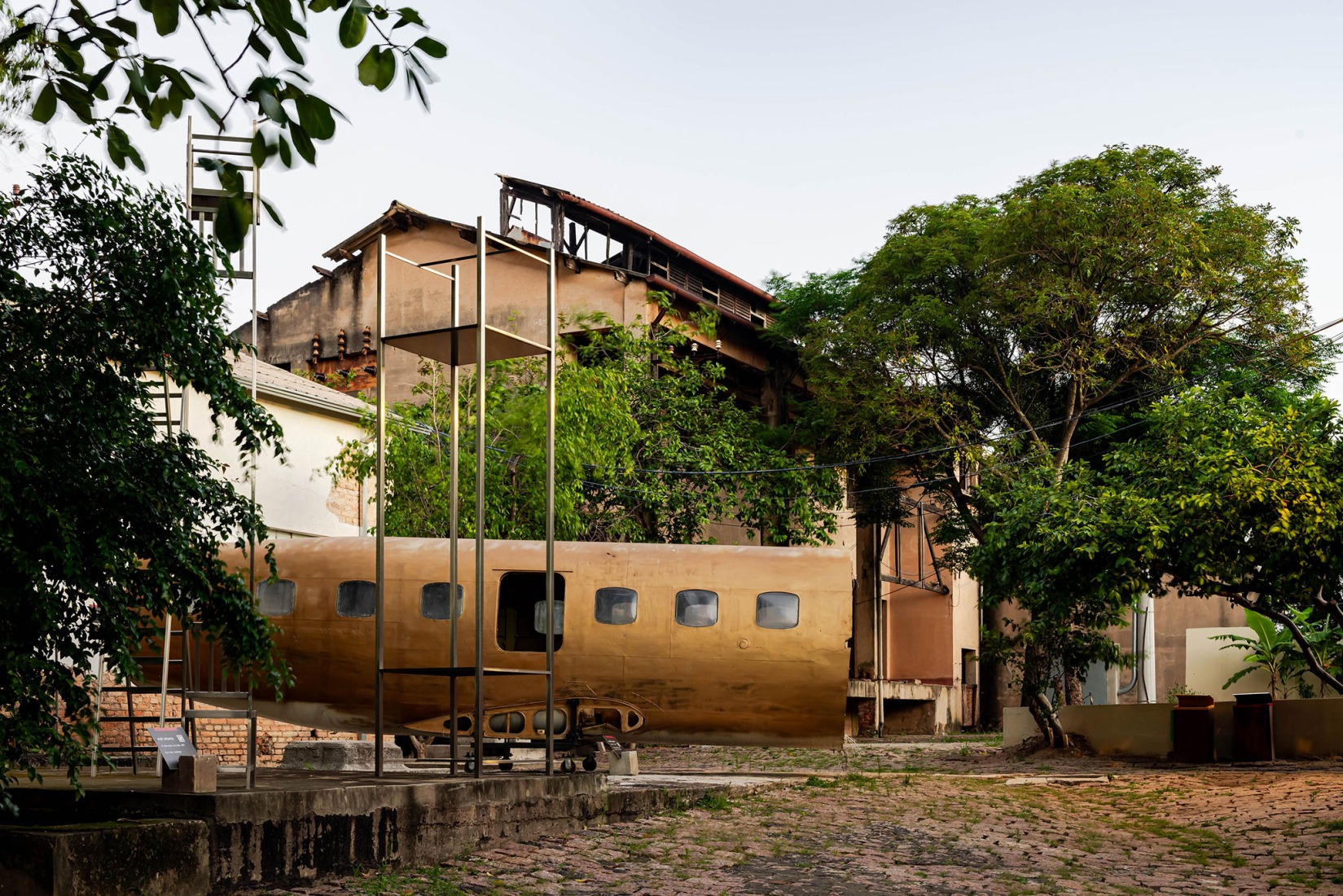  View of the corner between the Residential Atelier I, the Exhibition Building J, and the Atelier K, showcasing different renovation approaches within a green atmosphere and featuring a Marcos Amaro artwork. Fábrica de Arte Marcos Amaro (FAMA) by KAAN. Photograph by Sebastian van Damme.