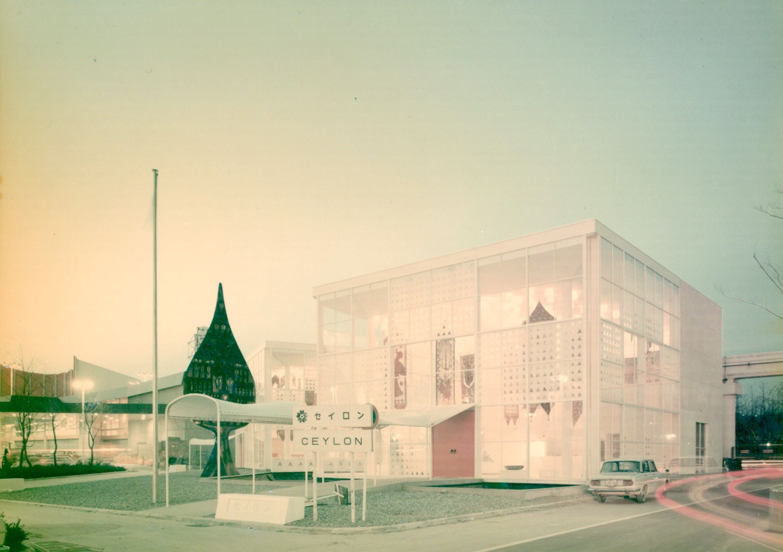 Exterior view of the two connected steel and glass cuboidal pavilions at Expo '70 with Laki Senanayake's Bo Leaf sculpture in the foreground, chromogenic colour print. Photograph by Geoffery Bawa (attributed), 1970. Courtesy by Geoffrey Bawa and Lunuganga Trusts, all rights reserved.