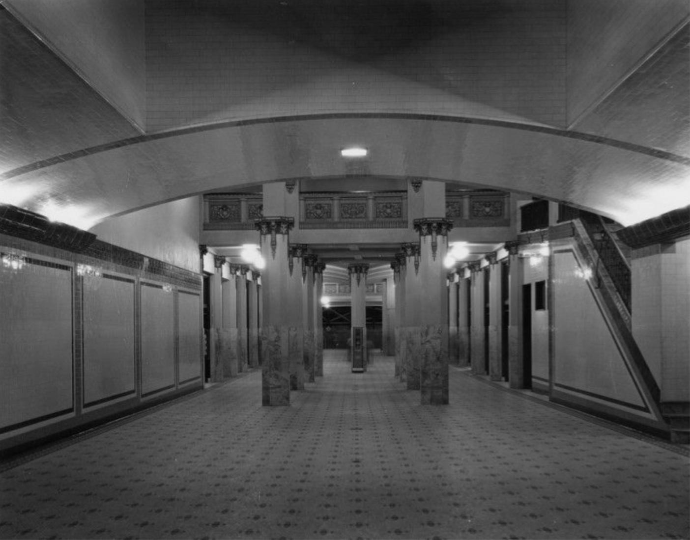 A view of Jergins Subway, a pedestrian walkway that ran under the busy intersection of Ocean Blvd. & Pine Ave. from 1927-1967. The tunnel ran under Ocean Boulevard and connected with the Jergins Arcade which was also a walk-through with a series of shops along the way and opened at the far end onto the Pike Amusement Park. The floor is paved with mosaic tiles and the walls with ceramic ones. The columns are capped with decorative friezes. The ceiling is slightly arched and a staircase is on the right. Los A