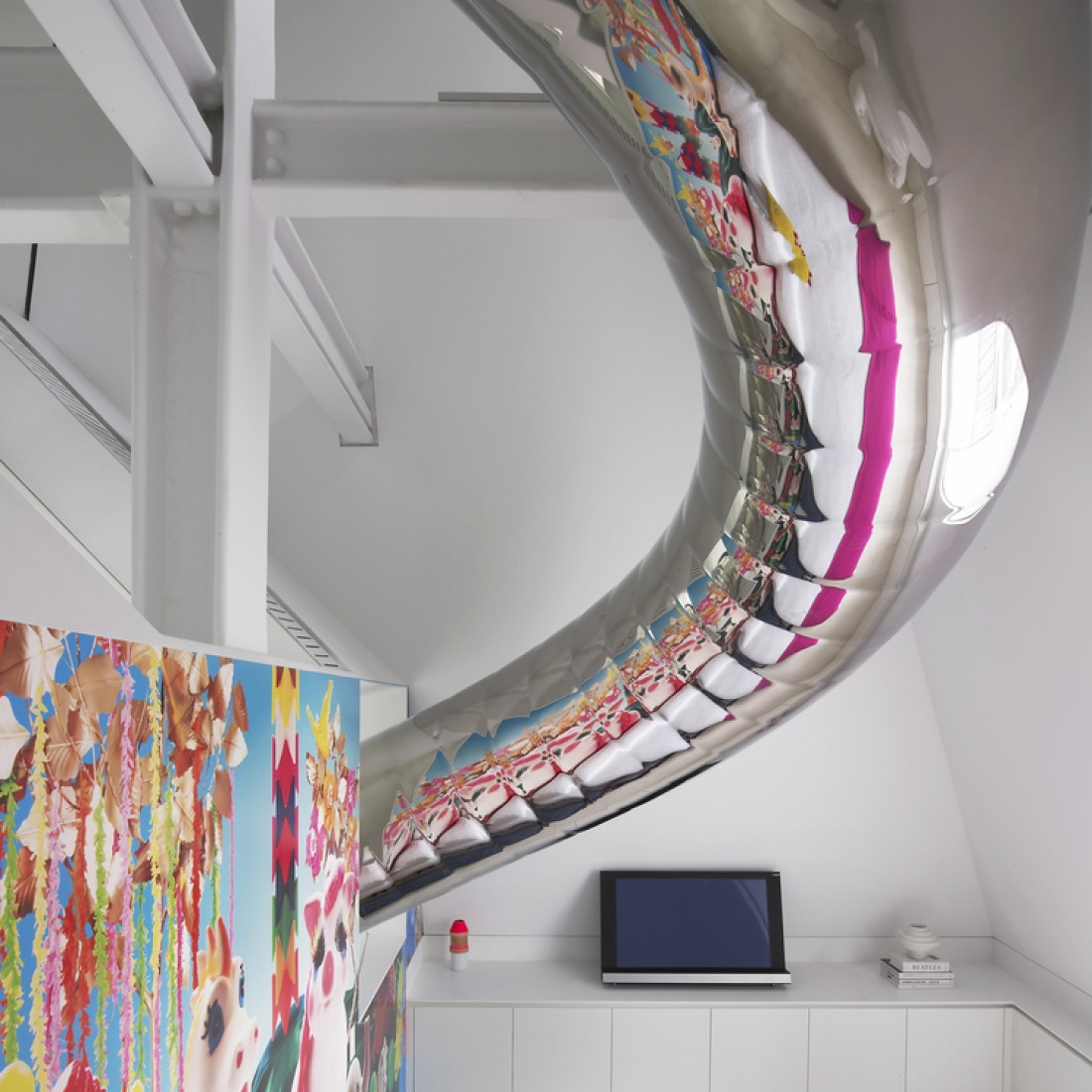 The cylindrical helical slide flares to an ellipse which is sectioned on the angle of the inclined glass wall  resulting in a circular opening where the slide emerges through the glass.  This circular opening creates an illusion of flatness contradicted by the sideways path of the slide as it begins its descent. 